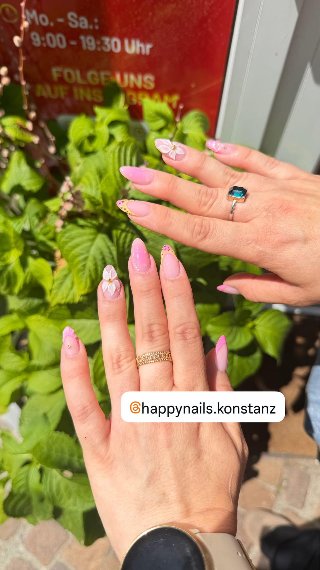 Elegante rosa Maniküre mit floralen Designs vor grünem Laub – Happy Nails Konstanz